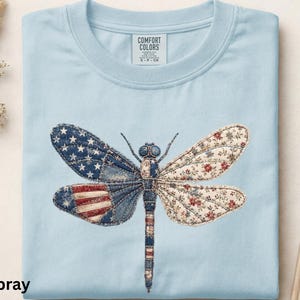Może przedstawiać: Jasnoniebieski t-shirt z grafiką ważki. Lewe skrzydła mają niebiesko-biały wzór w gwiazdki oraz czerwono-białe paski, podczas gdy prawe skrzydła mają kremowy kwiatowy nadruk. Tułów ważki posiada niebiesko-biały wzór.