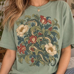 以下が含まれることがあります： セージグリーンのTシャツ。フロントには、ダークグリーンのヘビが、大きなクリーム色の花、赤いチューリップのような花、そして小さな赤い実をつけた複雑な緑の葉の間をうねるように描かれた、詳細なグラフィックプリントが施されています。