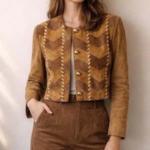 May include: A brown suede jacket with a chevron pattern and gold buttons. The jacket is paired with brown corduroy pants. The jacket has a round collar and long sleeves. The stitching is a lighter color than the jacket.