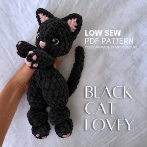 Può includere: Un peluche a forma di gatto lavorato all'uncinetto in nero con accenti rosa su orecchie e zampe, caratterizzato da grandi occhi scuri e un naso rosa. Il testo recita: LOW SEW PDF PATTERN YOU CAN MADE IN ANY COLOUR! BLACK CAT LOVEY. Ha una lunga coda.