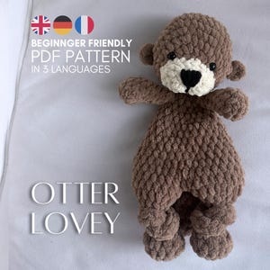 Könnte beinhalten: Ein braunes Häkel-Otter-Kuscheltier mit cremefarbenem Gesicht sowie schwarzer Nase und Augen. Der Text lautet: BEGINNGER FRIENDLY PDF PATTERN IN 3 LANGUAGES OTTER LOVEY. Das Stofftier hat eine strukturierte, weiche Garnoptik und kleine Ohren.