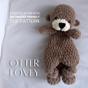 May include: A brown and cream-coloured crocheted otter lovey with black safety eyes and a black nose. The text on the image reads: ENGLISH & FRENCH BEGINNGER FRIENDLY PDF PATTERN OTTER LOVEY. The plush toy features a textured, soft yarn finish.