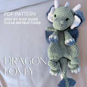 May include: A handmade crochet dragon lovey in shades of green, blue, and white. The dragon features a detailed design with wings, horns, and a tail. The image also includes text that reads "PDF PATTERN STEP-BY-STEP GUIDE CLEAR INSTRUCTIONS" and "DRAGON LOVEY".