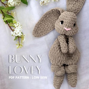 Könnte beinhalten: Ein gehäkelter Hasen-Schmusetuch aus dickem, hellbraunem Garn mit langen Ohren, schwarzen Sicherheitsaugen und einer kleinen, rosa gestickten Nase. Der Text BUNNY LOVEY PDF PATTERN - LOW SEW steht in Weiß auf der linken Seite.
