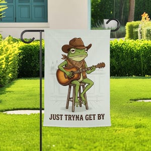 May include: A rectangular garden flag featuring a green frog wearing a brown cowboy hat and a patterned neckerchief, sitting on a wooden stool while playing an acoustic guitar. The text "JUST TRYNA GET BY" is printed in brown at the bottom.