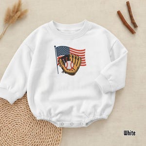 May include: A white long-sleeve baby bodysuit featuring a central embroidered graphic of a brown baseball glove holding a white baseball with red stitching, positioned in front of a waving American flag with red, white, and blue colours.