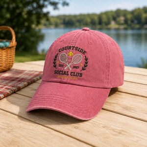 May include: A faded red baseball cap featuring an embroidered design of two crossed tennis rackets, a yellow tennis ball, and the text "COURTSIDE SOCIAL CLUB EST. 1987 RACQUET SPORT" in dark green and yellow thread on the front panel.