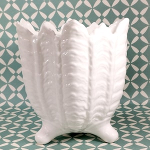 Może przedstawiać: Biała ceramiczna doniczka z falistym wzorem liści i trzema nóżkami.