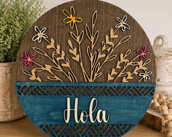 Decorazione floreale da appendere alla porta, realizzata a mano con strati di legno, dettagli color verde acqua e scritta "Hola" in rilievo.