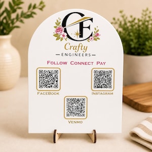 May include: A white arched sign with the words "Crafty Engineers" and social media QR codes for Facebook, Instagram, and Venmo. The sign has a floral design and is supported by a wooden stand.