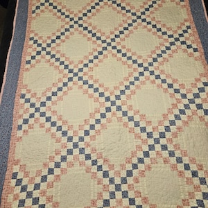 May include: A rectangular quilt featuring a repeating double wedding ring pattern. The design consists of cream-colored fabric sections framed by a grid of small, alternating navy blue and pink floral squares, finished with a navy blue border.