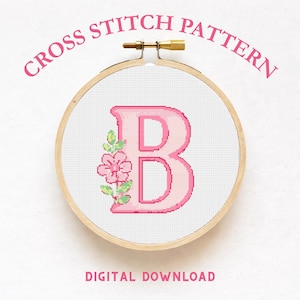 May include: A cross stitch pattern featuring a large, light pink letter B centered on white fabric, decorated with a small pink flower and green leaves on the left side. The design is held in a round wooden embroidery hoop with a metal tightening screw.
