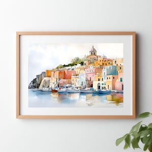 May include: Watercolor painting of a coastal town with colorful buildings in shades of orange, pink, and blue. White boats are docked in the water, reflecting the town's colors. The artwork is framed in a light brown wooden frame.