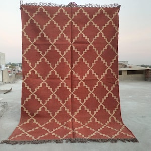 May include: A large, rectangular rug with a geometric pattern. The rug features a terracotta-colored background with a repeating diamond lattice design in a lighter beige hue. The edges are finished with a fringe.