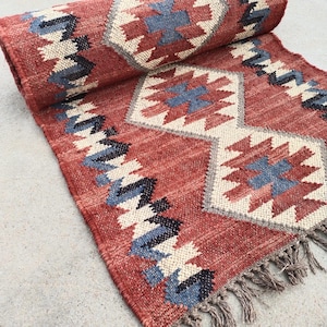 May include: A rolled-up red rug with geometric patterns in blue, cream, and black. The rug has a fringed edge and a textured surface, showcasing a traditional design. The rug is suitable for home decor.
