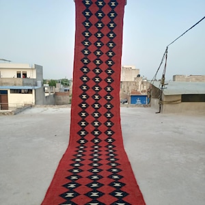 Puede incluir: Una alfombra pasillera larga y rectangular con un fondo rojo vibrante y un patrón repetitivo de rombos negros. Cada rombo negro contiene una pequeña forma de reloj de arena blanca centrada. La alfombra tiene flecos cortos en ambos extremos.