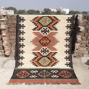 May include: A rectangular rug with a cream-colored background and a central geometric pattern in orange, black, and green. The rug has a dark brown border and fringe along the bottom edge. The rug is made of a woven material.