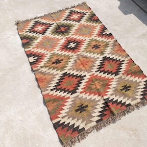 May include: A rectangular rug with a geometric pattern in shades of red, orange, black, green, and cream. The rug has a fringed edge and is likely made of woven material. The design features repeating diamond shapes.