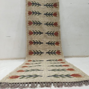 May include: Beige jute runner rug with a floral pattern in red, green, and navy blue. The rug features a repeating design of stylized flowers and leaves. The edges are finished with a fringe.