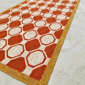 Könnte beinhalten: Ein rechteckiger Teppich mit einem sich wiederholenden geometrischen Muster aus orangefarbenen Achtecken, die durch weiße Linien getrennt sind. Der Teppich ist mit einer einfarbigen senfgelben Bordüre versehen, die das Design umrahmt.