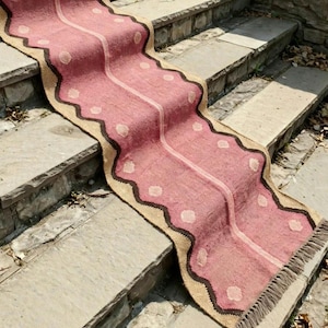 May include: A long, rectangular runner rug featuring a dusty rose pink center with a lighter pink vertical stripe. The rug has a wavy dark brown border and a natural tan edge, decorated with small, light-colored circular polka dot patterns.