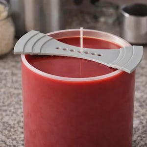Puede incluir: Una vela de pilar roja con una herramienta de centrado de mecha gris. La herramienta tiene una serie de agujeros para centrar la mecha y bordes curvos. La vela es cilíndrica y tiene una superficie lisa.