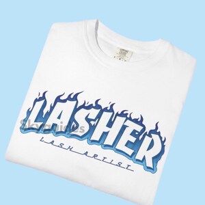 May include: White t-shirt with the word "LASHER" in a blue flame design, with "LASH ARTIST" below. The shirt is folded and displayed on a light blue background. The shirt has a crew neck and a tag.