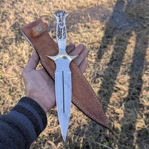 May include: A dagger with a polished steel blade and a handle made of bone and antler. The dagger has a brass guard and is accompanied by a brown leather sheath with a snap closure. The blade is approximately 10 inches long.