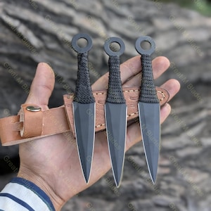 Puede incluir: Tres cuchillos arrojadizos negros con un anillo en la parte superior y mangos envueltos se muestran en una funda de cuero marr&oacute;n. Los cuchillos tienen una hoja afilada y puntiaguda. El fondo es una superficie de madera borrosa.