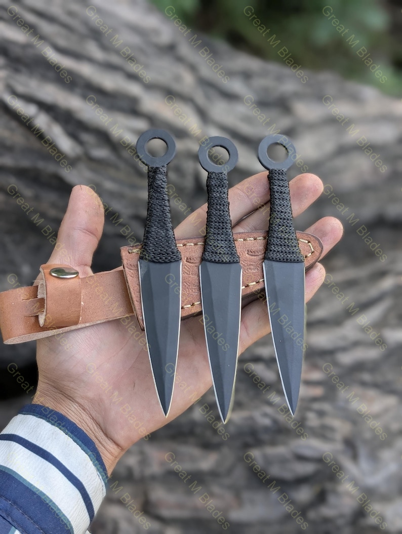 Puede incluir: Tres cuchillos arrojadizos negros con un anillo en la parte superior y mangos envueltos se muestran en una funda de cuero marr&oacute;n. Los cuchillos tienen una hoja afilada y puntiaguda. El fondo es una superficie de madera borrosa.
