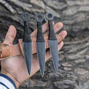 Puede incluir: Se muestran tres cuchillos arrojadizos negros con mangos tejidos y pomos anulares. Las hojas se guardan en una funda de cuero marr&oacute;n. El texto "Great M Blades" es visible en el fondo.