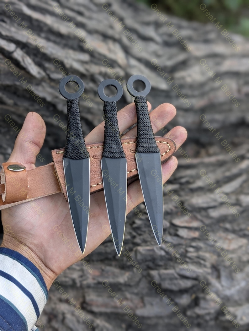Puede incluir: Se muestran tres cuchillos arrojadizos negros con mangos tejidos y pomos anulares. Las hojas se guardan en una funda de cuero marr&oacute;n. El texto "Great M Blades" es visible en el fondo.