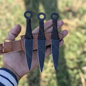 Puede incluir: Se muestran tres cuchillos arrojadizos negros con un mango de anillo y un agarre envuelto. Las cuchillas se sostienen en una mano, con una funda de cuero marr&oacute;n. Los cuchillos tienen un dise&ntilde;o elegante y puntiagudo.