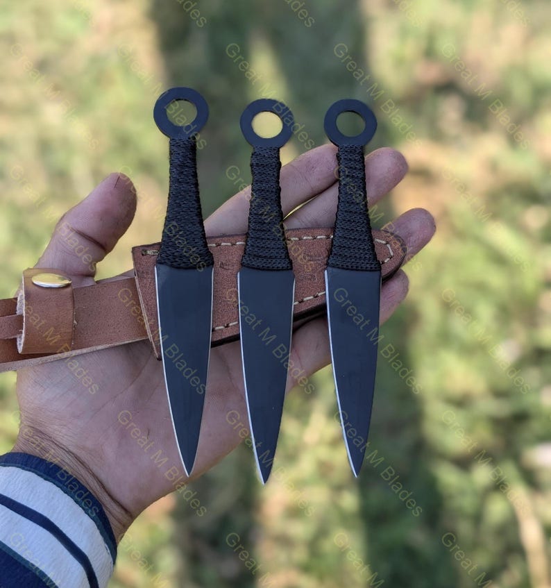 Puede incluir: Se muestran tres cuchillos arrojadizos negros con un mango de anillo y un agarre envuelto. Las cuchillas se sostienen en una mano, con una funda de cuero marr&oacute;n. Los cuchillos tienen un dise&ntilde;o elegante y puntiagudo.