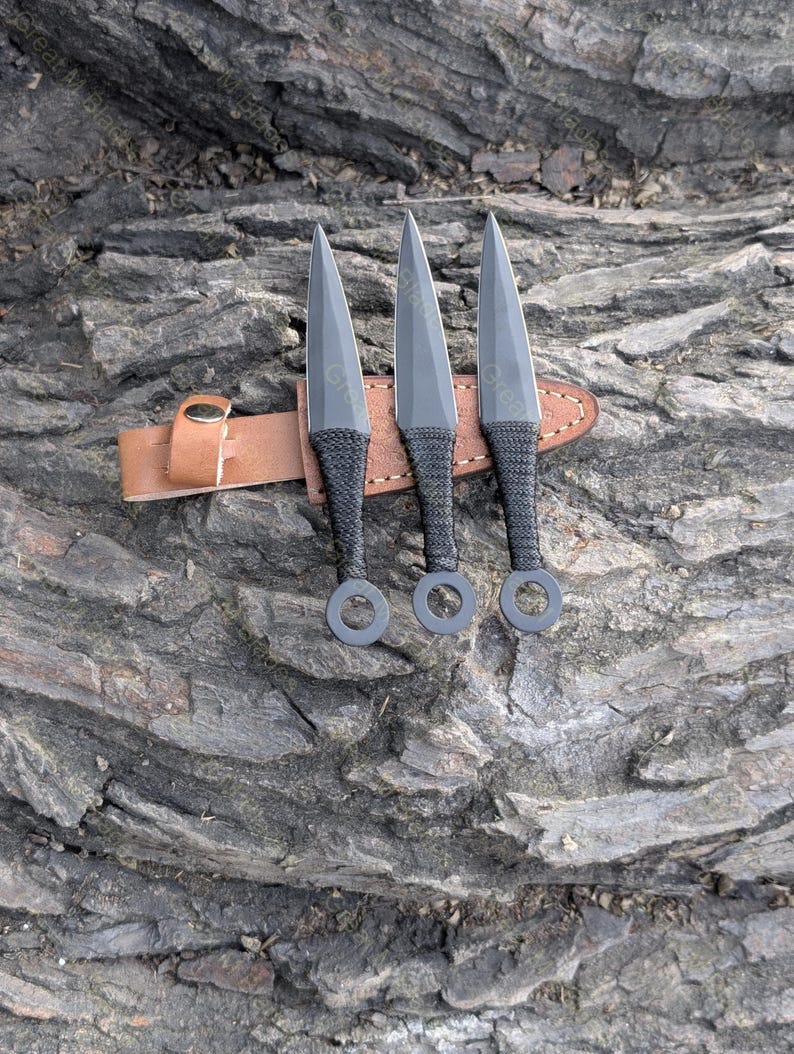 Puede incluir: Tres cuchillos arrojadizos negros con hojas puntiagudas y mangos envueltos descansan sobre una funda de cuero marr&oacute;n. Los cuchillos tienen un anillo en la base del mango. La funda est&aacute; asegurada con un cierre met&aacute;lico. Fondo texturizado marr&oacute;n oscuro.