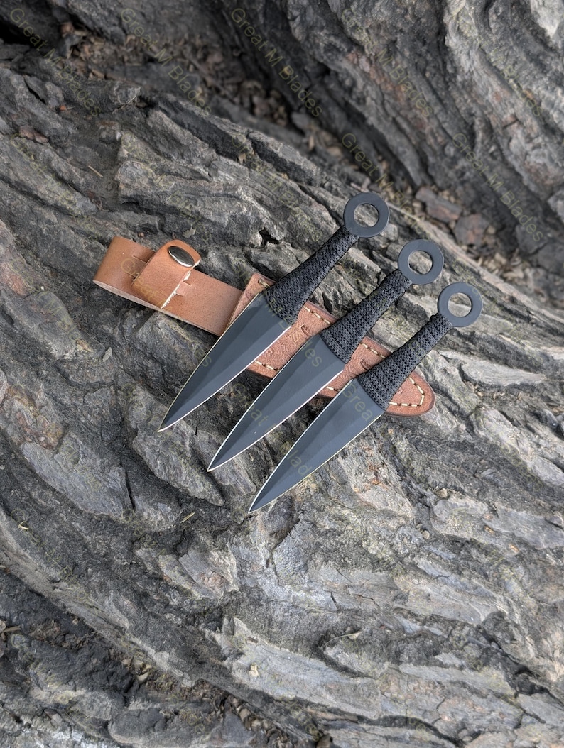 Puede incluir: Tres cuchillos de lanzamiento negros con una funda de cuero. Los cuchillos tienen una hoja puntiaguda y un anillo en el extremo del mango. La funda es marr&oacute;n y tiene un cierre de bot&oacute;n a presi&oacute;n. El texto "Great M Blades" es visible en los cuchillos.