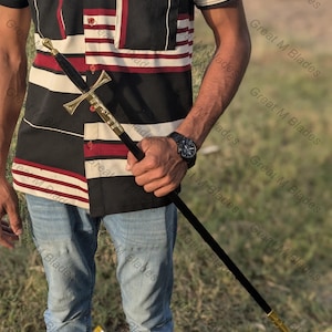 May include: A long, slender sword featuring a black scabbard with gold-toned metal accents at the hilt and tip. The hilt has a cross-shaped guard and a rounded pommel. The sword is held in a vertical position, showcasing its sleek design and metallic details.
