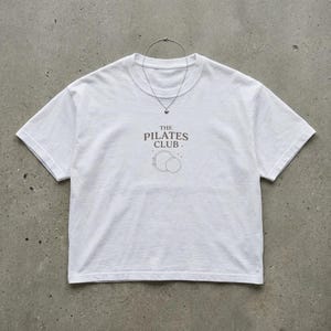 May include: A white t-shirt featuring the text "THE PILATES CLUB" in brown, and a graphic of two circles. A silver necklace with a small pendant is draped on the shirt. The shirt is made of a soft material.