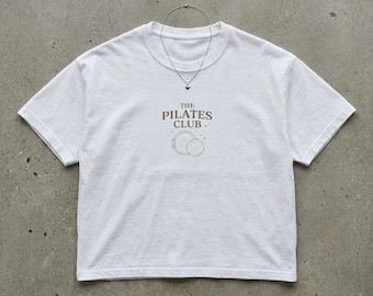 Puede incluir: Una camiseta blanca con el texto "THE PILATES CLUB" en marrón, junto con un gráfico de dos círculos. Un collar plateado con un pequeño colgante cuelga de la camiseta. La camiseta es de un material suave.