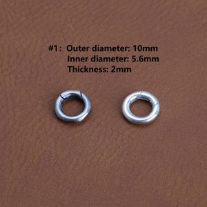 Può includere: Due anelli metallici circolari color argento con una fessura. Il testo indica che il diametro esterno è di 10 mm, il diametro interno è di 5,6 mm e lo spessore è di 2 mm. Gli anelli sono su una superficie marrone.