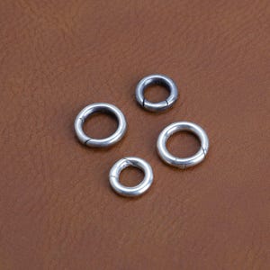 Può includere: Quattro piccoli anelli metallici color argento con un design a cerniera sono disposti su una superficie marrone. Gli anelli sono circolari, con un piccolo spazio che indica la cerniera. Il metallo ha una superficie leggermente riflettente.