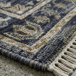 May include: A rectangular area rug featuring a woven texture in shades of navy blue and beige. The rug displays a repeating geometric pattern with a decorative border and is finished with short, off-white fringe tassels along the edge.