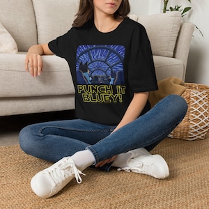 May include: A black short-sleeved t-shirt featuring a graphic of two blue dog characters in a spaceship cockpit looking out at stars streaking by. Below the graphic, the text reads PUNCH IT BLUEY! in yellow block letters on the black fabric.