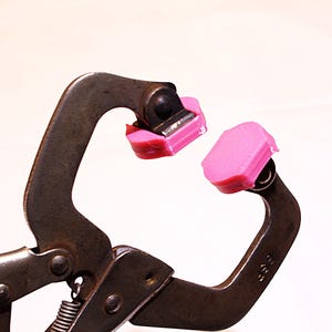May include: Close-up of a metal clamp with pink rubber pads on the jaws. The clamp is open, revealing the spring and hinge mechanism. The pads are a bright pink color.