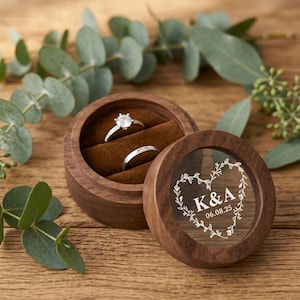May include: A round wooden ring box with a brown velvet interior holding two silver rings. The lid features a clear circular window engraved with a heart-shaped vine design, the letters K & A, and the date 06.08.25 in white lettering.