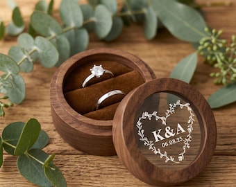 Könnte beinhalten: Eine runde Ringbox aus Holz mit braunem Samtinnenfutter und zwei Silberringen. Der Deckel hat ein klares, rundes Sichtfenster, das mit einem herzförmigen Rankenmuster, den Buchstaben K & A sowie dem Datum 06.08.25 in Weiß graviert ist.