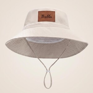 May include: A beige bucket hat featuring a brown rectangular leather-like patch on the front with the name Bella printed in black script. The hat includes a white mesh interior lining and a matching beige chin strap with an adjustable toggle.
