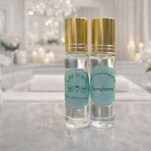 May include: Two clear glass perfume bottles with gold caps and light blue labels. One label reads "Liv & Viv" with three small animal illustrations. The other label reads "Symphony". The bottles are on a reflective surface.