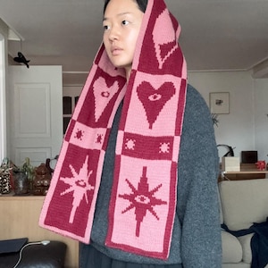May include: A knitted scarf with a hood, featuring a repeating pattern of hearts, stars, and geometric shapes in shades of pink and burgundy. The scarf is worn over a gray sweater.