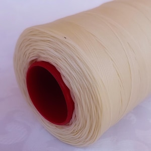 May include: A spool of off-white thread with a red core. The thread is tightly wound, showing a textured surface. Ideal for sewing, embroidery, or other crafts.
